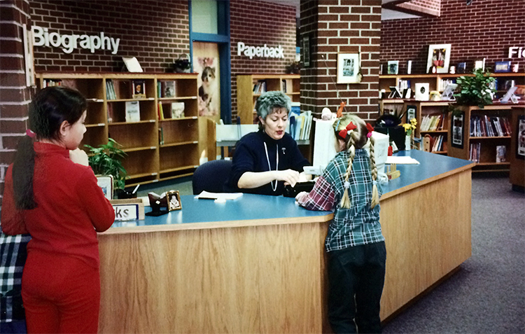 Photograph of two students checking out library books. Library Assistant Theresa Werner is seated at the long check-out desk, and the students have their backs to the camera. The library has dark brown brick walls. There is a brick pillar next to the desk. Rows of bookshelves are visible in the distance, arranged into biography, paperback, and fiction sections. 