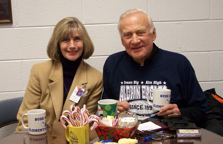 Photograph of Assistant Principal Barbara Gist with Dr. Buzz Aldrin. They are seated at a small table that has food in the center. Dr. Aldrin is wearing an Aldrin Eagles shirt.