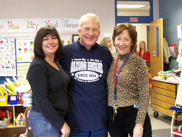 Photograph of teachers Mrs. Harrison and Mrs. Keyser posing with Dr. Buzz Aldrin. They are standing in a classroom and two adults and a child are visible in the doorway behind them.