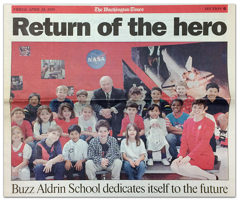 Newspaper clipping of the cover of the Washington Times newspaper from April 28, 1995. The headline reads: Return of the Hero, Buzz Aldrin School dedicates itself to the future. A large color photograph is beneath the headline. It shows Buzz Aldrin seated with a group of 20 students and their teacher on metal risers. On the wall behind them are photographs of a Space Shuttle, children at computers, and the NASA logo.