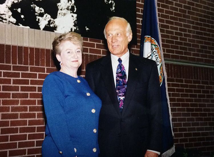 Photograph of Aldrin Elementary School Principal Gina Ross and Buzz Aldrin. They are standing side-by-side on the small raised platform inside the school lobby. A Virginia flag is visible behind Aldrin. 