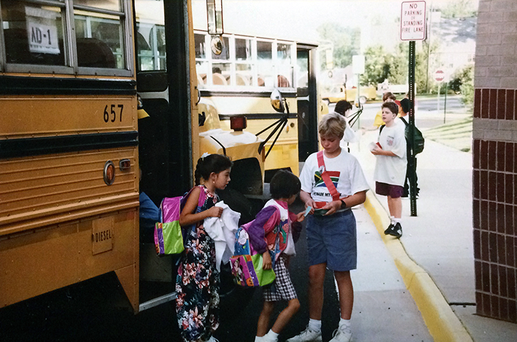 Photograph of students arriving at Aldrin Elementary School taken in 1994. Several school buses are parked along the sidewalk. Students are exiting the buses and heading into the building.
