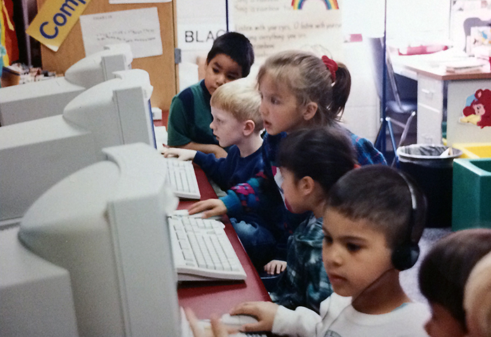 Photograph of students working at computers in a classroom. The picture was taken in 1994. Five children are pictured, working at three computers. One boy is wearing headphones.