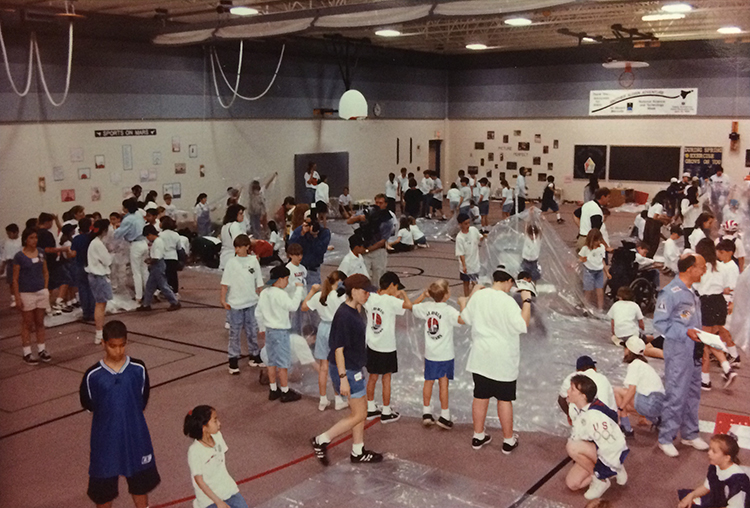 Photograph showing a large portion of Aldrin Elementary School's gymnasium during the Marsville project. Students are arranged in groups spread throughout the facility. They are working with large sheets of plastic, assembling structures. The initial stages of construction are pictured.