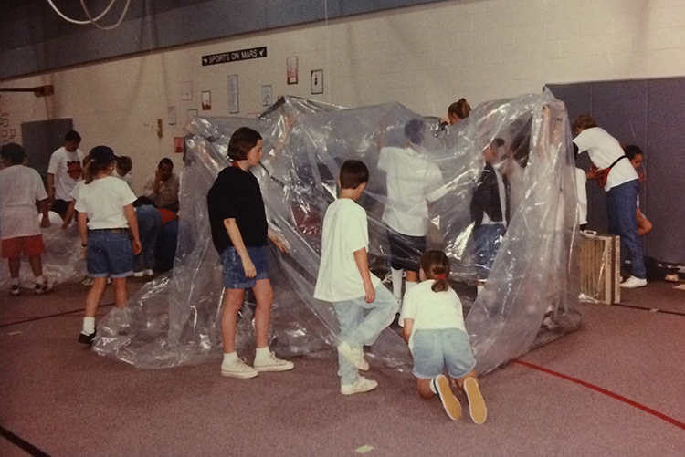 Close up photograph of students working on the Marsville Project in the gymnasium. A group of at least eight students is constructing a habitable structure out of a large sheet of see-through plastic. Several students are holding up the plastic so braces can be put in place to create a tent-like structure.