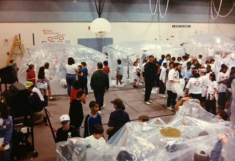 Photograph showing a large portion of the Aldrin Elementary School gymnasium during the Marsville project. At least three groups of students have finished constructing their plastic bubble tent-like habitats. In the foreground, two more are still being constructed. 