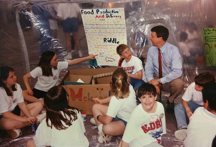 Photograph taken inside a completed Marsville Project bubble habitat. A group of eight students and an adult are pictured. The students are seated on the floor. A cardboard box, with the word Welcome written on it in several colors, is placed next to one of the plastic walls. Above the box, affixed to the wall, is a student-created poster entitled Food Production and Delivery. One of the students is pointing out a feature of the poster to the adult. 