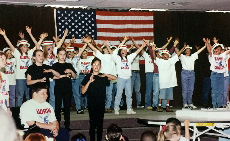 Photograph of students performing a musical at the Mission to Planet Earth Exhibition. A large group of students is standing on a stage. The children are singing in unison with their arms raised in the air. A large American flag is affixed to a black curtain behind them. The students are wearing white hats with red and blue trim, and white t-shirts printed with the words Aldrin Eagles and an illustration of an eagle in flight. In the foreground, three students in black are performing the musical with American Sign Language.  