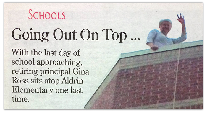 Newspaper clipping showing a photograph of Principal Ross atop the roof of Aldrin Elementary School. She is looking down over the edge, waving at the camera. Text next to the picture reads: Schools. Going Out On Top... With the last day of school approaching, retiring Principal Gina Ross sits atop Aldrin Elementary one last time.