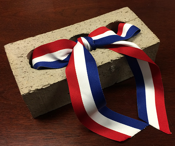 Photograph of a brick that was purchased for the exterior veneer of Aldrin Elementary School. A red, white, and blue ribbon has been threaded through the interior holes and tied in a bow. The brick is light gray and brown in color. Some surplus bricks were used as fundraising gifts. 