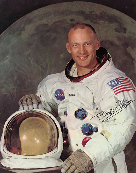 A light-skinned man with crew cut hair, smiling. He wears a white space suit, and rests his right hand on the helmet. The spacesuit has hose connectors on the front, along with a NASA logo next to 'Aldrin', which is embroidered on the top center of the suit. There is a large U.S. flag on the left shoulder. The helmet's transparent faceplate is tinted gold. The background is the Moon, tinted dark.