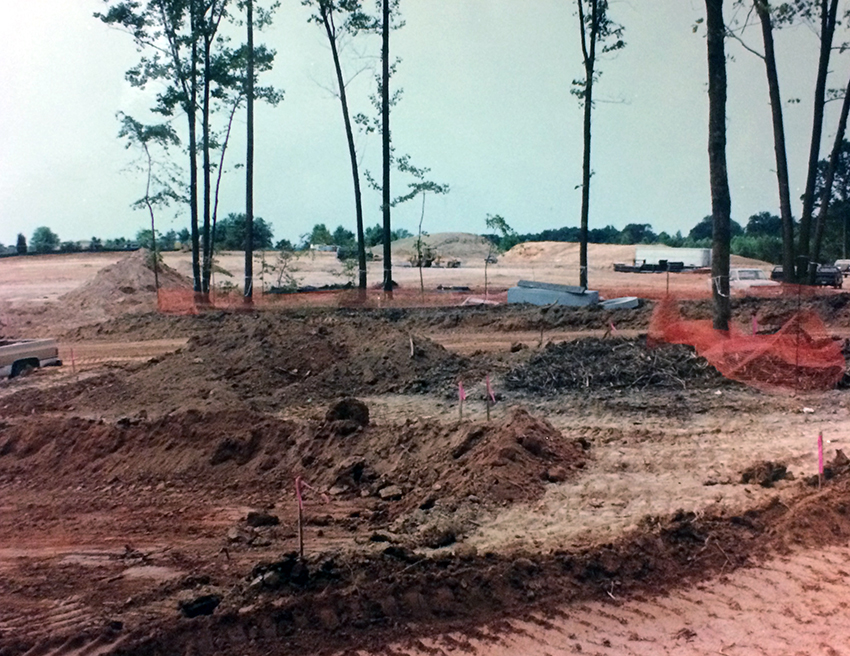 Photograph of the Aldrin site. The grounds have been cleared of trees and vegetation, with the exception of a few trees in the foreground. Heavy construction equipment can be seen in the distance, leveling the ground. The reddish brown dirt in the foreground has been churned up by the large tires of construction equipment.