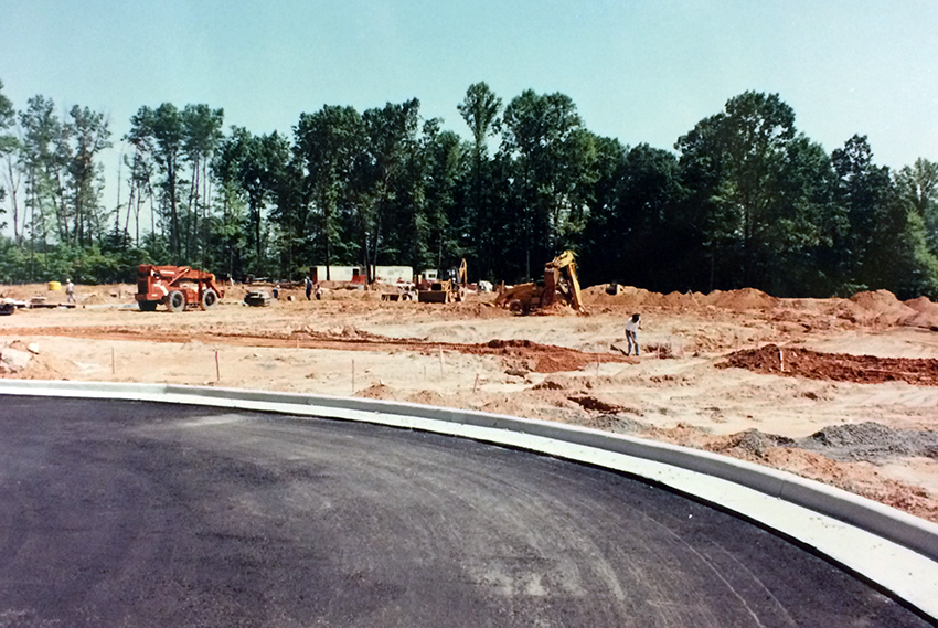 Photograph of the Aldrin site. Work is beginning on digging the foundation of the building. Two backhoes are visible in the distance. A construction worker walks along the edge of a trench in the mid-ground. A paved roadway with a concrete curb are visible in the foreground.