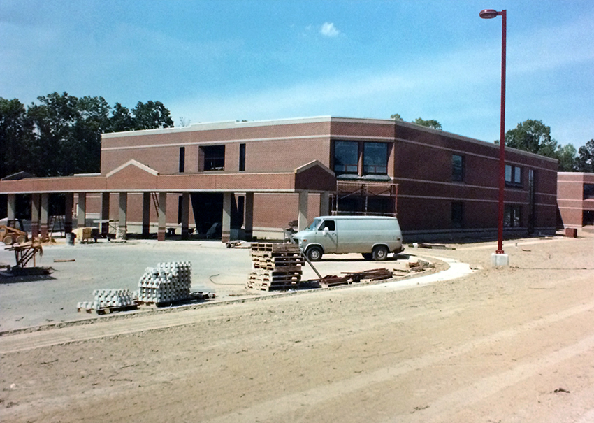 Photograph of the rear of Aldrin Elementary School taken from the south. Only one wing of the building is visible. The exterior looks complete with windows in place. A driveway with concrete curb are visible on the left and a construction van is parked close to the building. Empty wooden pallets have been stacked on the driveway. The school grounds have been graded, but the grounds closest to the building have not been sodded.