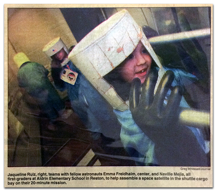 Newspaper clipping from a Fairfax Journal article in 1998, showing three students participating in a space simulation. The children are wearing blue full-body suits with large black rubber gloves, and have white buckets on their heads with a window opening for their faces. The article caption reads: Jacqueline Ruiz, right, teams with fellow astronauts Emma Freidhaim, center, and Naville Mejia, all first-graders at Aldrin Elementary School in Reston, to help assemble a space satellite in the shuttle cargo bay on their 20-minute mission.