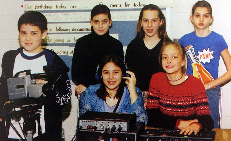 Yearbook photograph of Aldrin Elementary School’s news team from the 2001 to 2002 school year. Six students are pictured, five girls and one boy. The boy is operating a VHS camcorder on a tripod, and two girls are sitting at a table with audio and video technical equipment in front of them. Three girls stand behind the table against the wall.