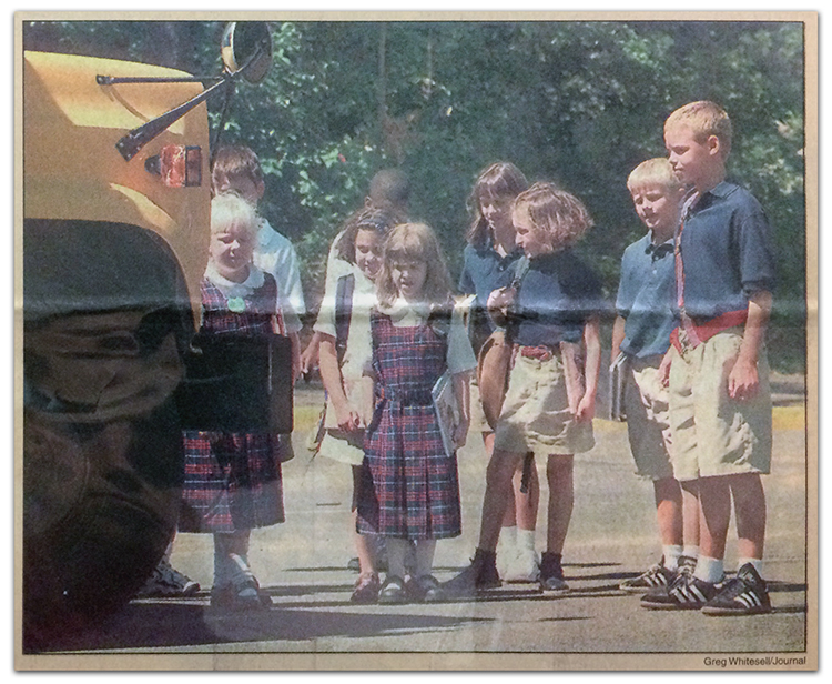 Photograph of a newspaper article clipping from The Journal about the uniform policy at Aldrin Elementary School. The clipping is a color photograph of a group of nine Aldrin students waiting to board a school bus. Two girls are wearing the plaid jumper school uniform, and the remaining students are wearing the khaki and navy blue uniform. 